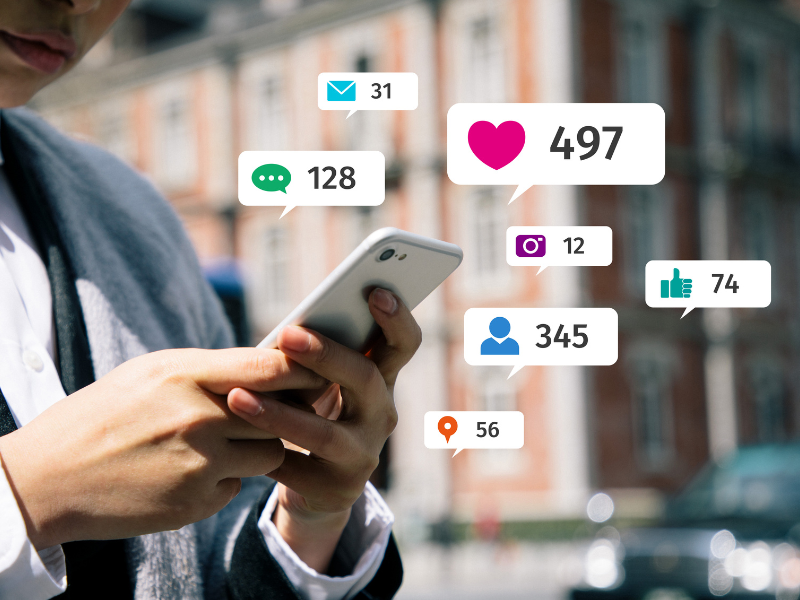 The image displays someone using a smart phone with icons emerging from the phone including likes, hearts, comments, friends, chats, and mail.