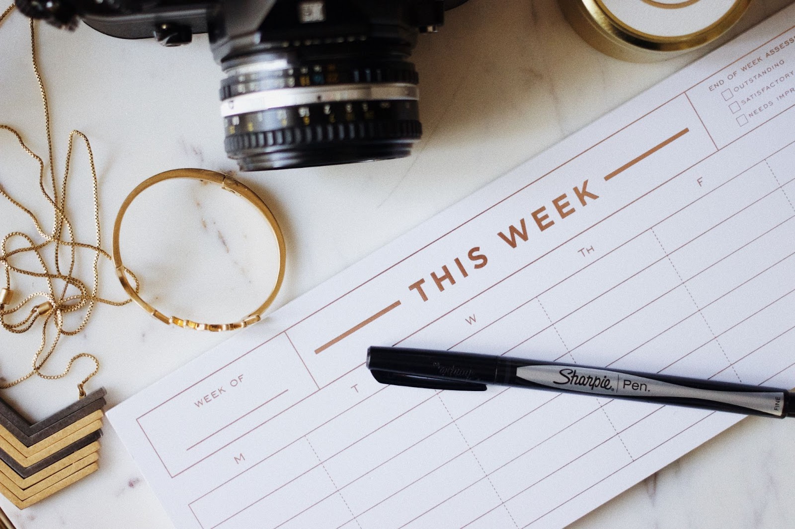 A calendar on a white marble desk surrounded by a camera, jewelry, and a Sharpie pen.