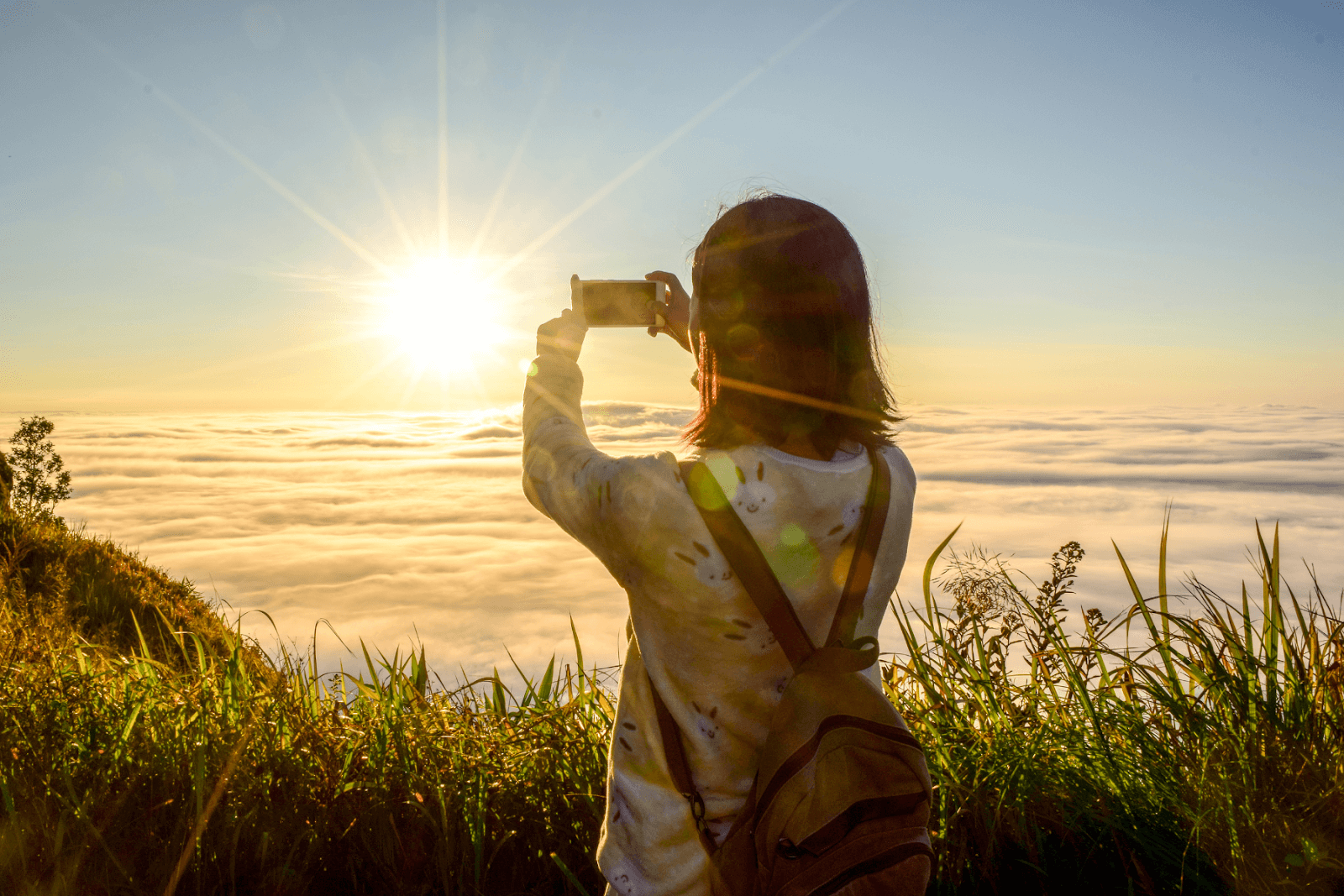 The photo displays a woman taking a photo of a sunset high above the clouds. She is surrounded by green grass on a hilltop as the sun plunges beneath the scattered clouds.