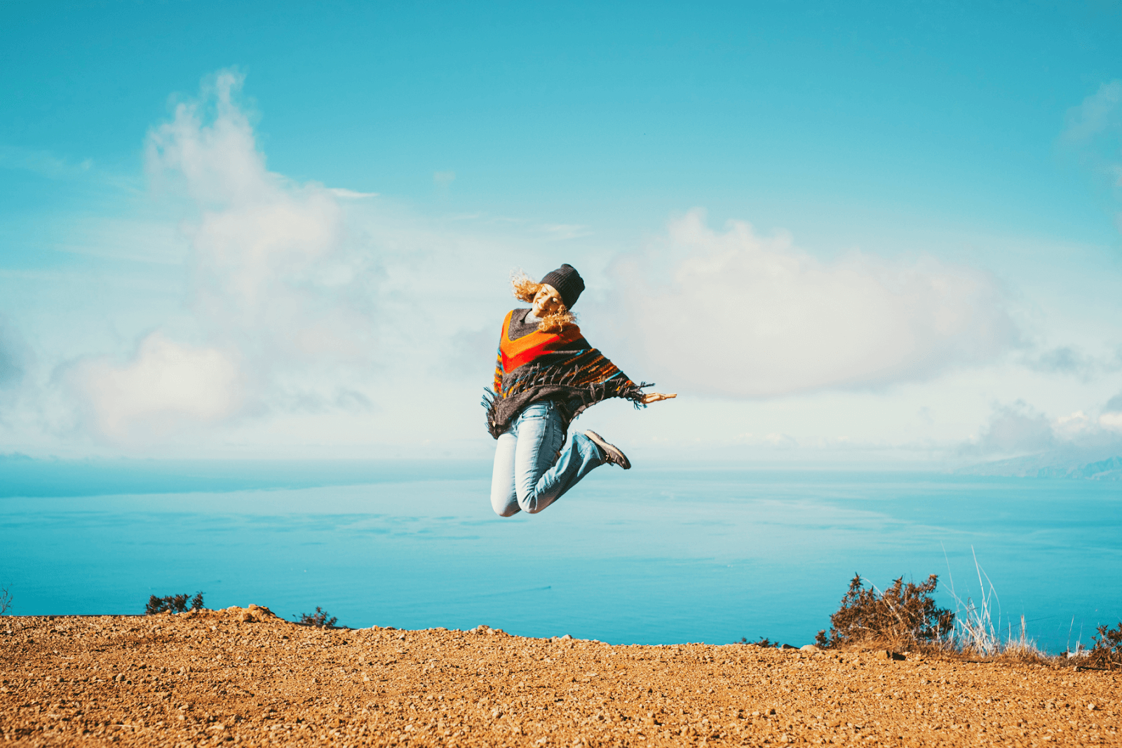 The image displays a woman jumping into the air on top of a mountain with a beautiful ocean in the background. The mountaintop is dirt with a few shrubs, while the woman wears a beanie, jeans, and a poncho.