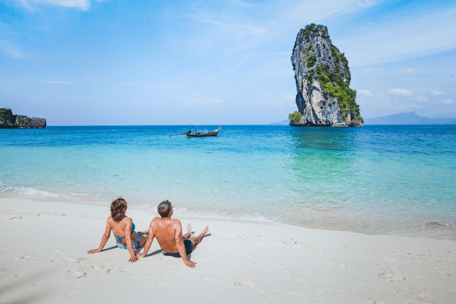 The image displays a couple relaxing on a beach in Asia. The beach has white sand with turquoise waters, and a large rock jets upward from the ocean floor with a small boat nearby.