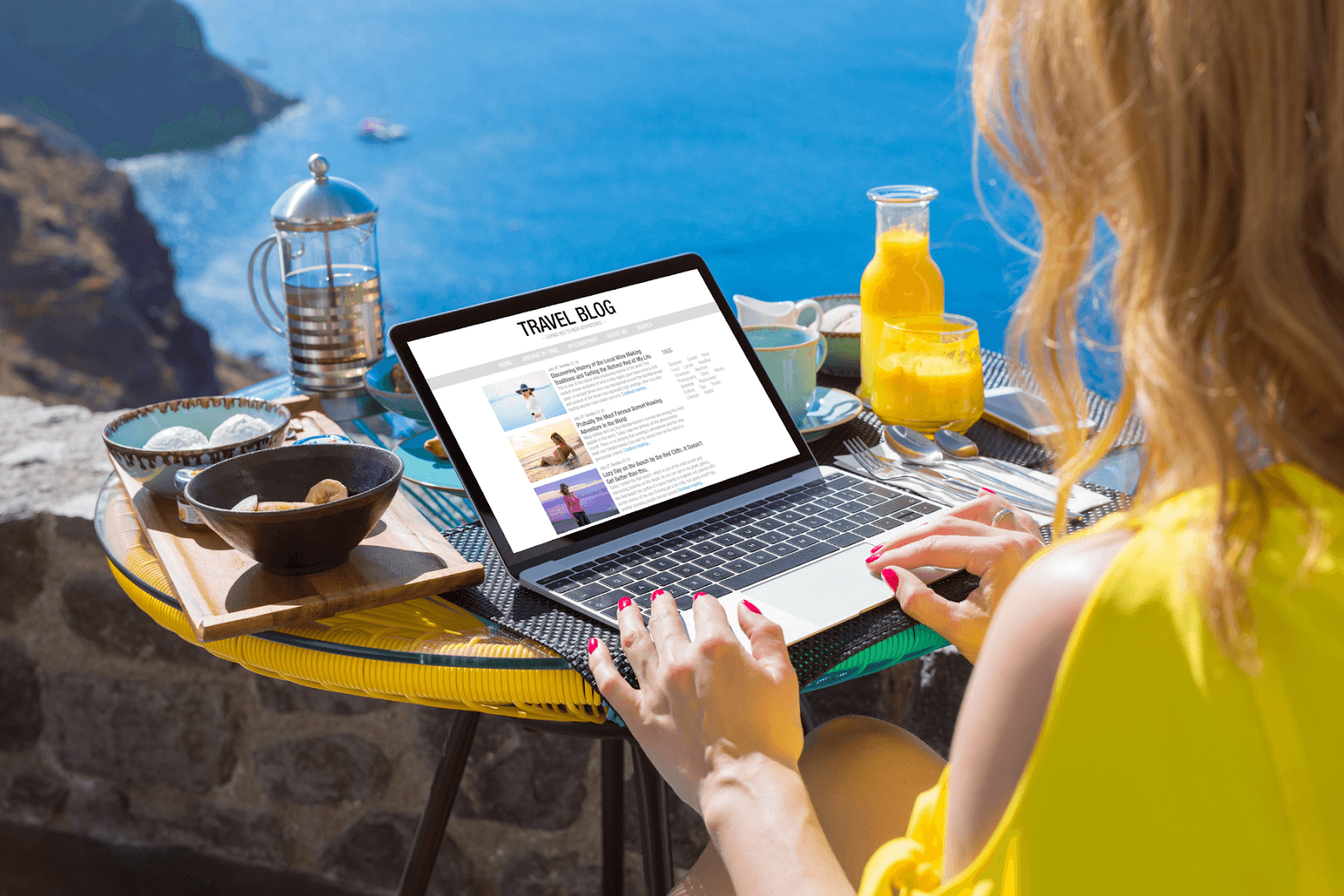 The photo displays a woman sitting at a small table with food, drinks, and a laptop. She is working on her travel blog as she overlooks a beautiful cliffside before plunging below into the ocean. A catamaran is in the distance floating on the water.